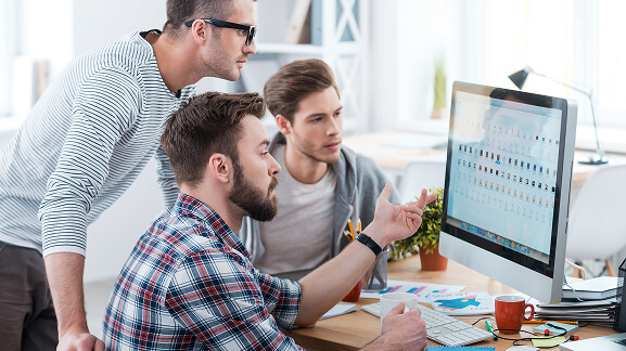 Three colleagues collaborating around a desktop monitor in a modern office.
